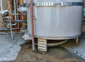 Close-up of an aging furnace or heating unit with rust stains, connected copper and metal pipes, and visible wear around the base. Water stains and dark patches cover the concrete floor beneath the equipment.